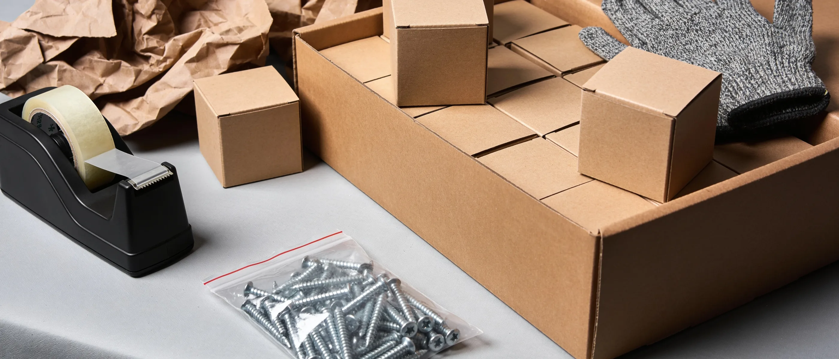 Offener Versandkarton mit Kleinkartons, Beutel Spanplattenschrauben und Arbeitshandschuh auf dem Packtisch
